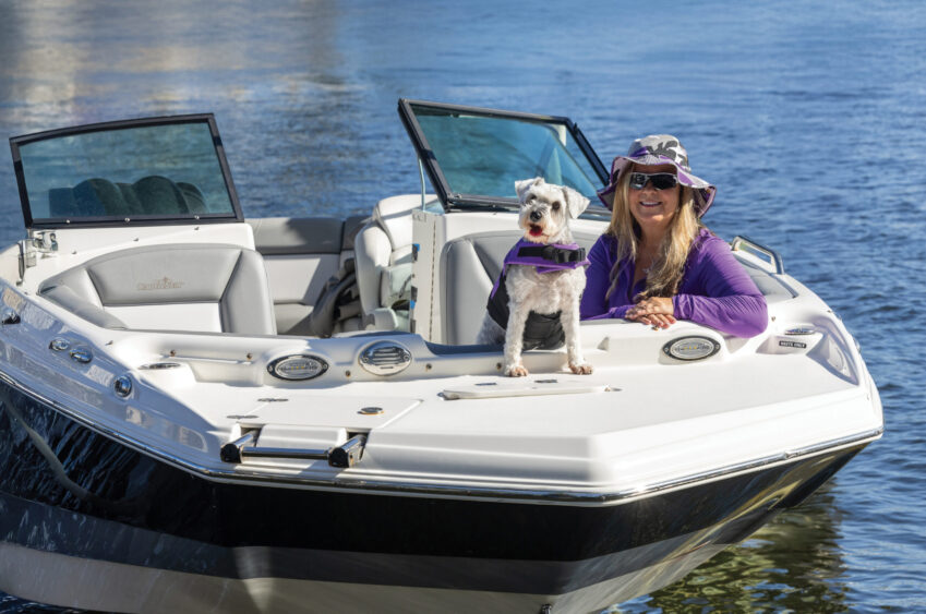 A woman in a purple shirt and hat sits with a small white dog in a life jacket on the bow of a white boat floating on calm water, perfectly capturing the Dog Days of Summer. Lifestyle