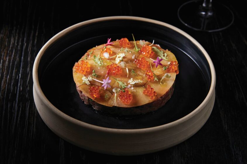 A round gourmet dish topped with orange fish roe, flower petals, and herbs sits on a black plate with a wooden rim, placed on a dark wooden table—a refined celebration of the vibrant Flavors of Miami. Lifestyle