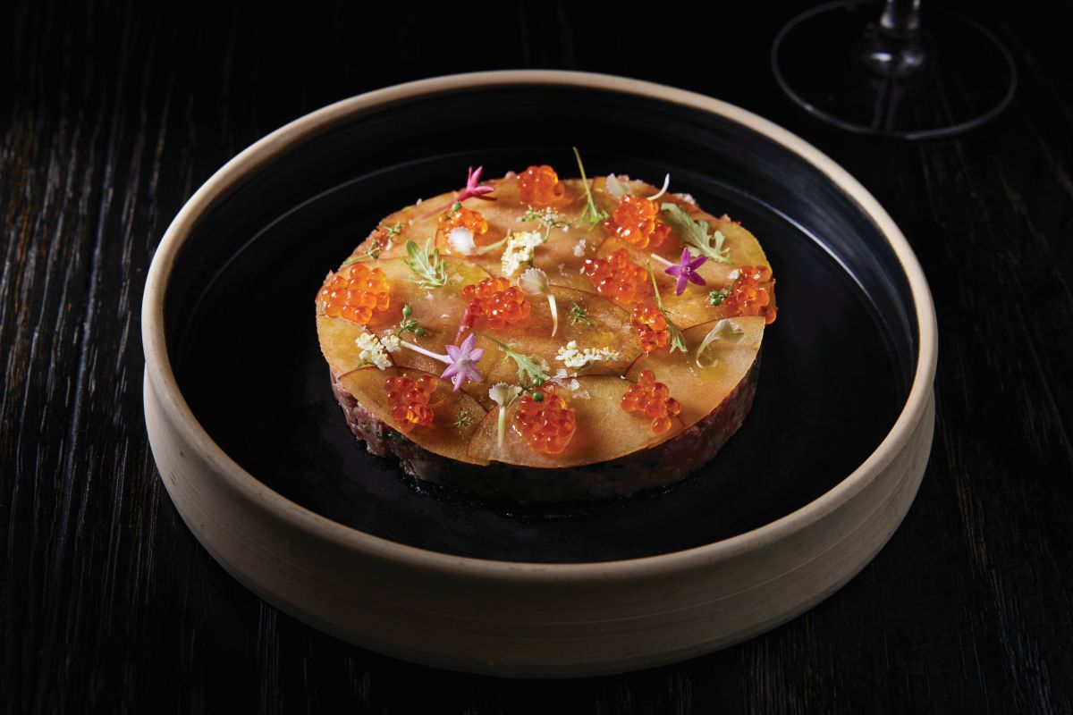 A round gourmet dish topped with orange fish roe, flower petals, and herbs sits on a black plate with a wooden rim, placed on a dark wooden table—a refined celebration of the vibrant Flavors of Miami. Lifestyle