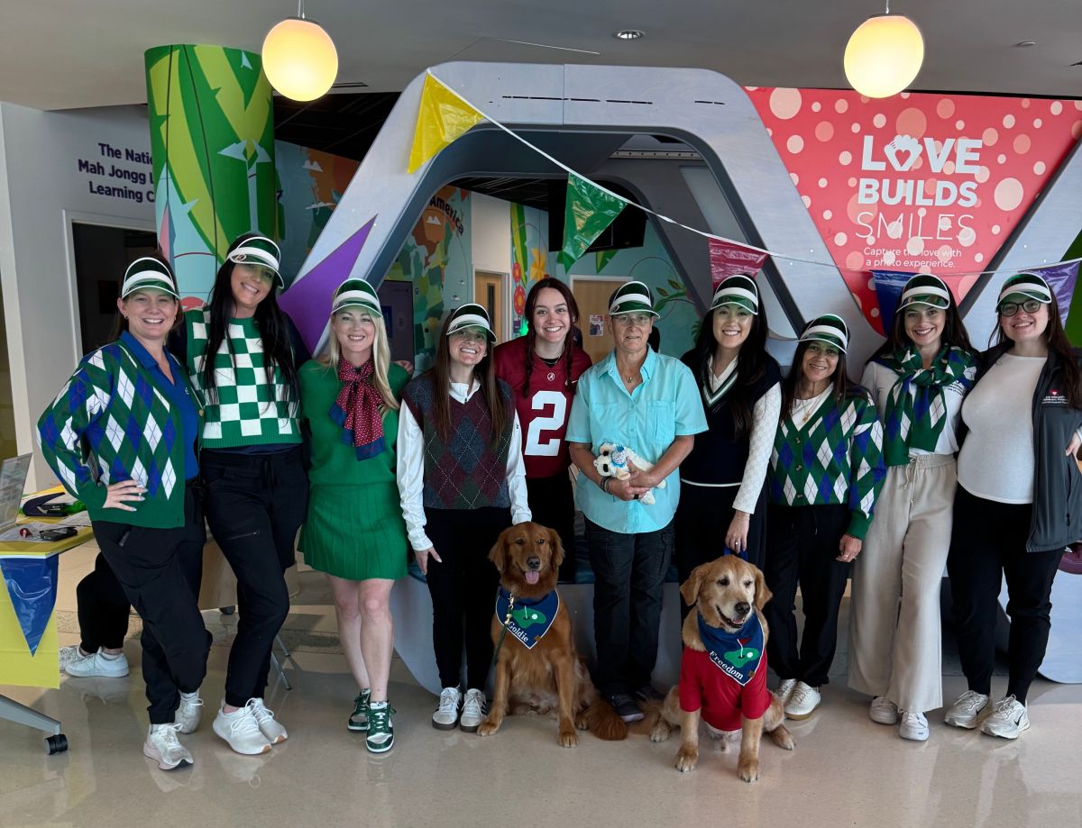 A group of nine smiling people in golf-themed outfits pose indoors with two golden retrievers wearing Paws With Purpose vests. Colorful banners and a "Love Builds Smiles" sign are in the background. Lifestyle