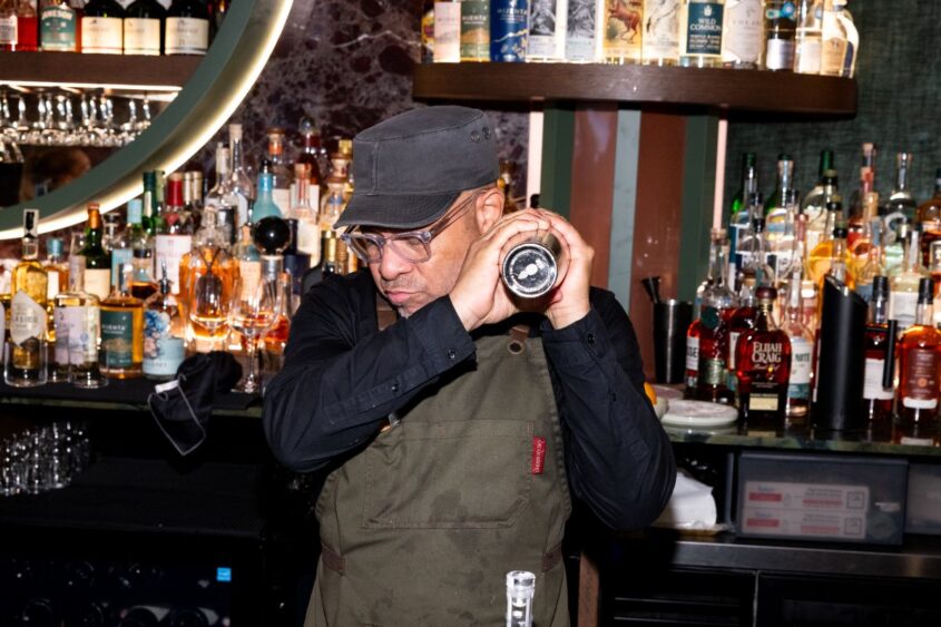 A bartender wearing glasses, a cap, and an olive apron shakes a cocktail shaker behind a bar stocked with various liquor bottles in South Miami’s popular Flight West. Lifestyle