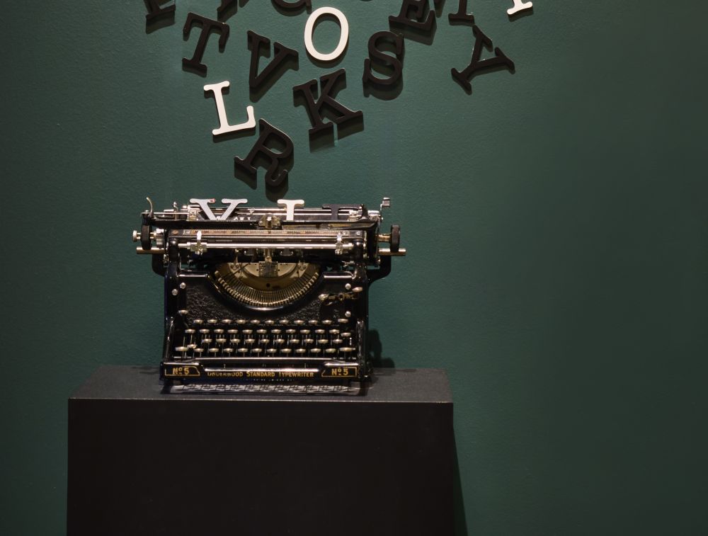 A vintage typewriter sits on a black pedestal, with scattered black and white letters suspended above it against a dark green background—evoking the timeless charm of the Grand Dame of Key West. Lifestyle