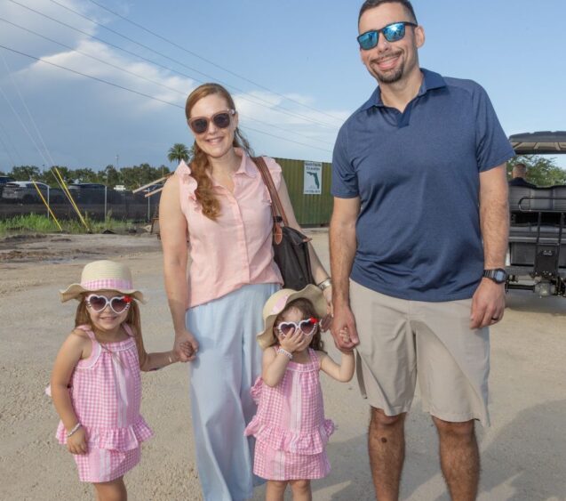 A smiling family of four, two adults and two young girls in matching pink outfits and heart sunglasses, stands outdoors on a sunny day near Ave Maria. Lifestyle