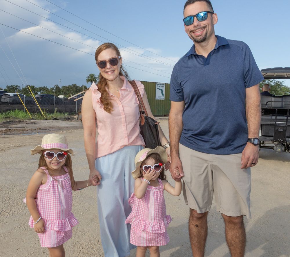A smiling family of four, two adults and two young girls in matching pink outfits and heart sunglasses, stands outdoors on a sunny day near Ave Maria. Lifestyle