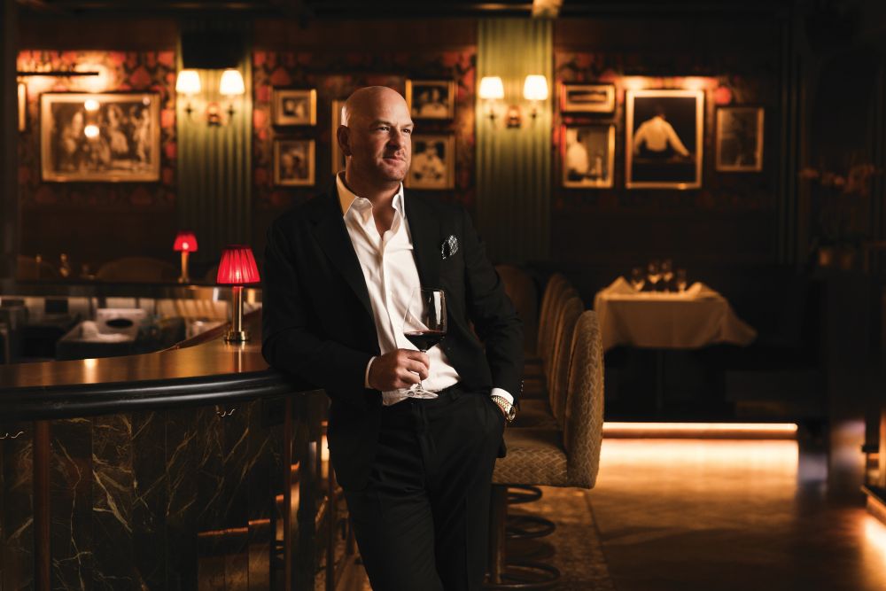 A man in a suit leans against a bar, holding a glass of wine in a dimly lit, luxury restaurant on Las Olas, with framed photos on the wall and warm ambient lighting, evoking the sophisticated atmosphere inspired by Marc Falsetto. Lifestyle