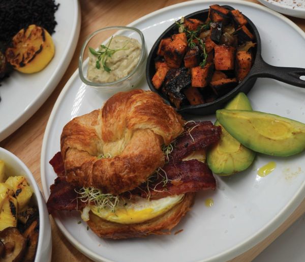 A croissant sandwich with bacon, egg, and sprouts, served with sliced avocado, a small bowl of sauce, and a skillet of roasted sweet potatoes and vegetables on a white plate—a delightful option inspired by airline wellness trends to elevate your in-flight dining experience. Lifestyle