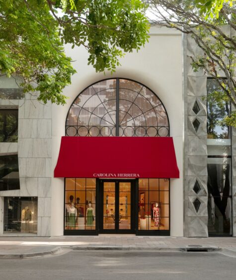 The Carolina Herrera store exterior in the Design District features a large arched window, a bright red awning with the brand name, and displays elegant clothing inside. Marble and modern buildings flank the boutique. Lifestyle
