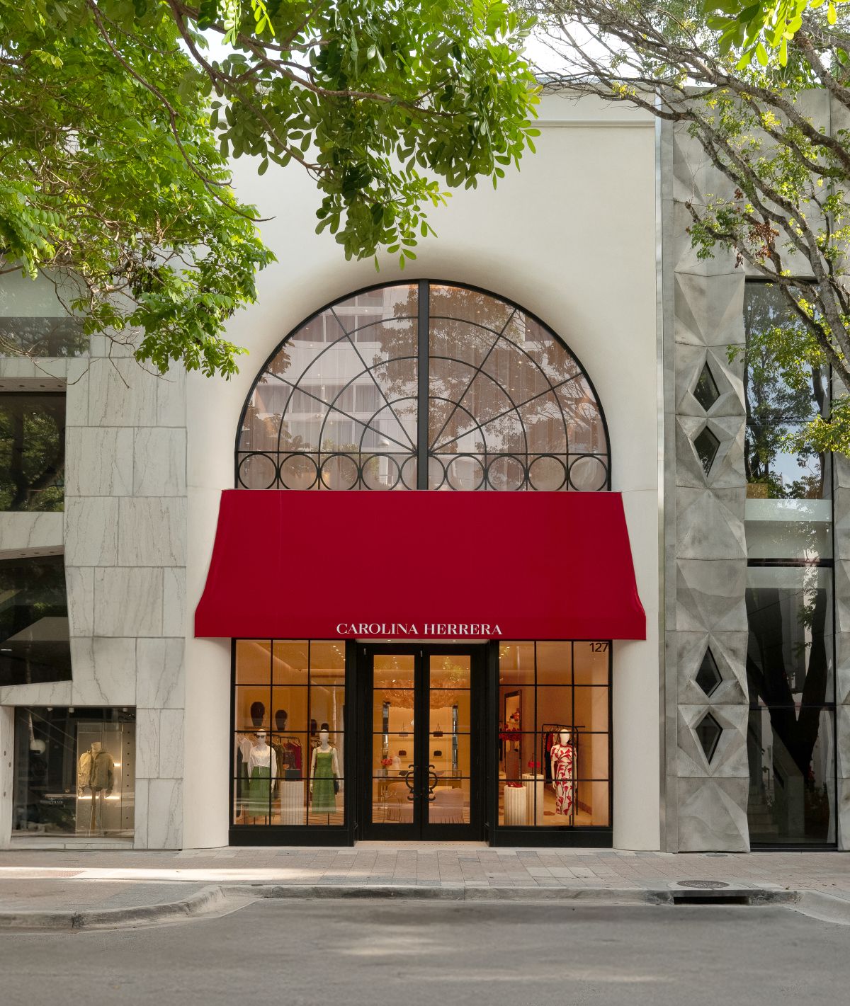 The Carolina Herrera store exterior in the Design District features a large arched window, a bright red awning with the brand name, and displays elegant clothing inside. Marble and modern buildings flank the boutique. Lifestyle