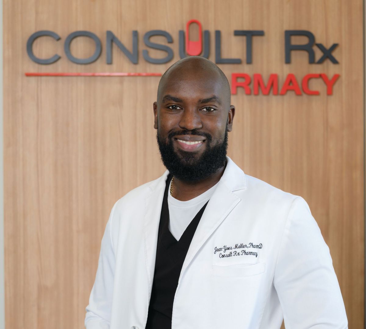 A smiling man in a white lab coat stands in front of a wooden wall with “CONSULT Rx PHARMACY” written on it, embodying the spirit of Flipping the Scrip through innovative pharmacy care. Lifestyle
