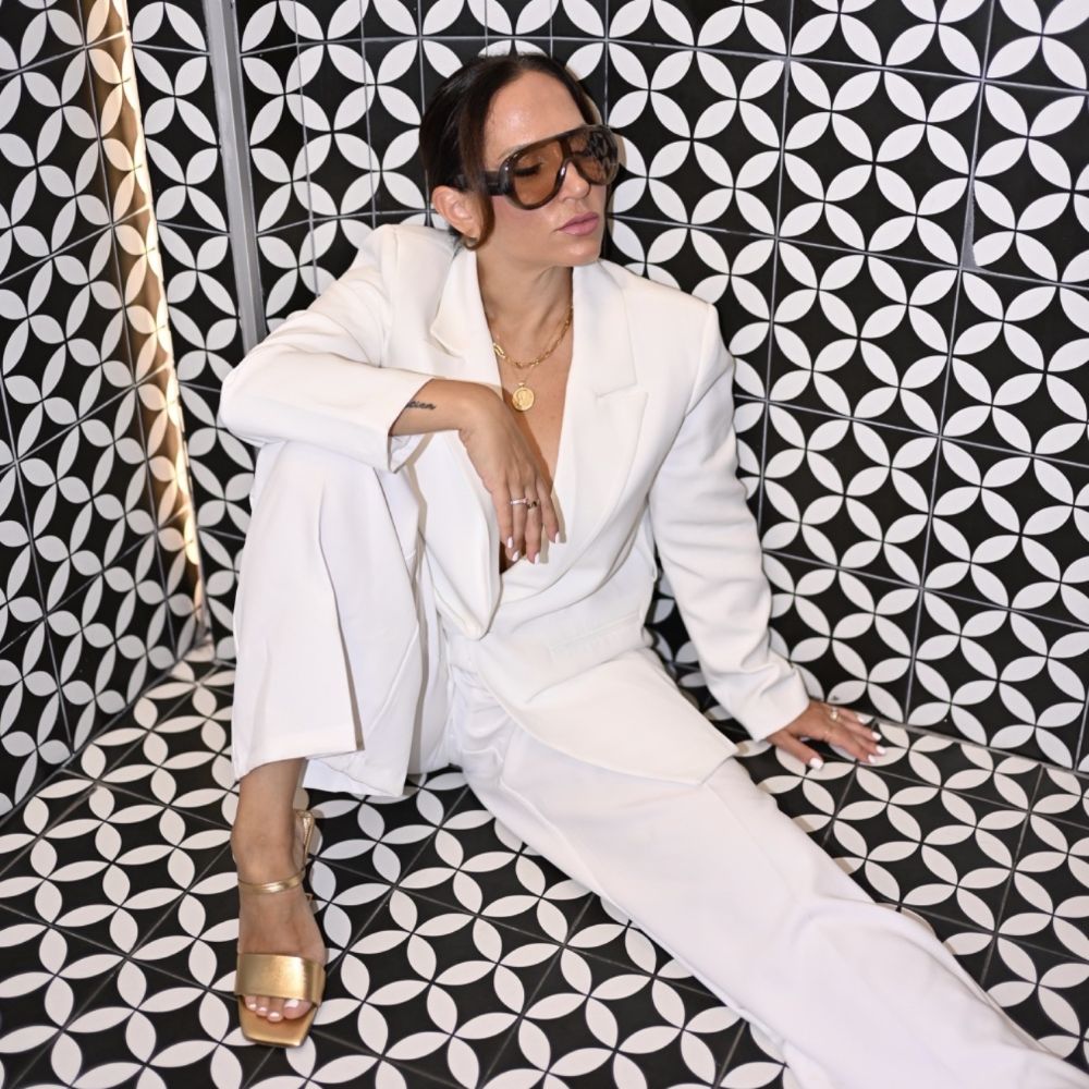 A woman in a white suit and gold sandals sits against a black-and-white geometric patterned wall, exuding confidence with her Pilates-inspired poise. She wears large sunglasses and layered gold jewelry, embodying the strength and elegance of the Pilates Powerhouse. Lifestyle