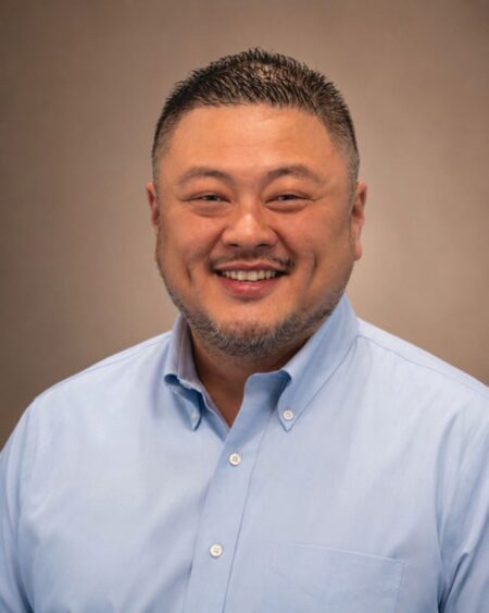 A smiling man with short dark hair and a trimmed beard, wearing a light blue button-up shirt, poses for a professional headshot against a plain, brownish background—confidently carrying the torch of professionalism and warmth in his demeanor. Lifestyle