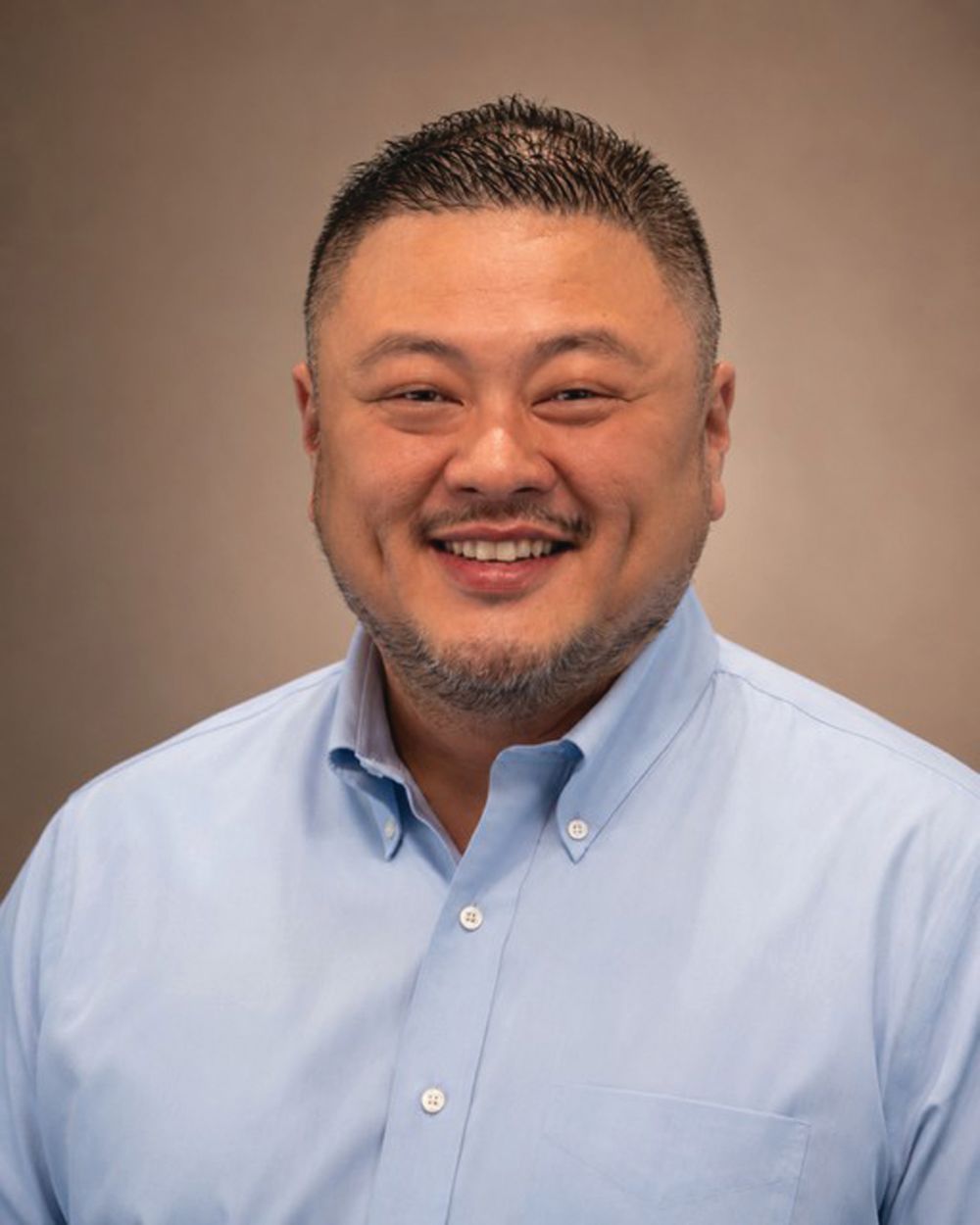 A smiling man with short dark hair and a trimmed beard, wearing a light blue button-up shirt, poses for a professional headshot against a plain, brownish background—confidently carrying the torch of professionalism and warmth in his demeanor. Lifestyle