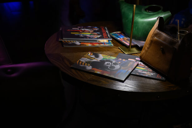A dimly lit table with magazines featuring colorful covers, a brown purse, and a green object, possibly a decorative watering can, under a lamp. Lifestyle
