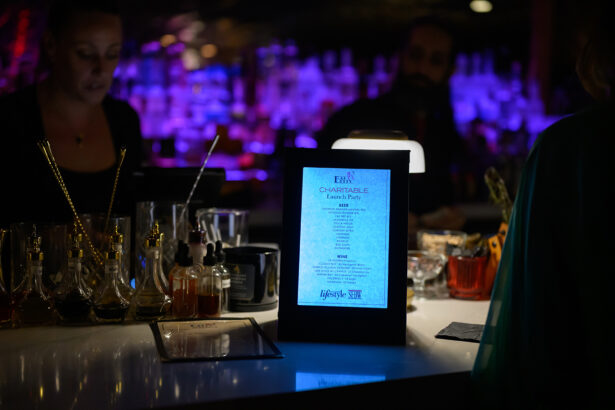 A dimly lit bar with bottles and glasses; a glowing menu in the center reads “Charitable Launch Party.” Two people stand behind the bar with a purple-lit background. Lifestyle