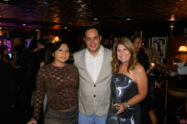 Three people posing and smiling at a dimly lit, stylish bar or lounge. Two women flank a man, all dressed in evening attire. Other guests are in the background, enjoying drinks and conversation. Lifestyle
