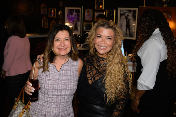 Two women smiling and posing together at an indoor event. One woman holds a drink. Other guests and framed photos can be seen in the background. Lifestyle