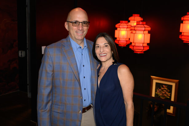 A smiling man in a blue plaid blazer and glasses stands next to a woman in a navy dress. Red lanterns hang in the dim background, and a framed picture is visible on the wall behind them. Lifestyle