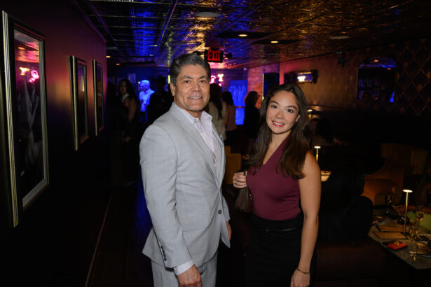 A man in a light gray suit and a woman in a burgundy top pose and smile in a dimly lit lounge with people socializing in the background. Lifestyle