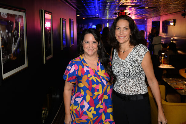 Two women smile and pose together in a dimly lit, modern lounge with framed photos on the wall and blue and pink lights in the background. Lifestyle
