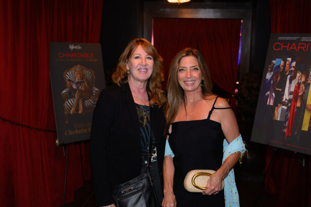 Two women smiling and posing together at an event, standing in front of red curtains and movie posters. One holds a clutch, the other a black purse. Both wear dresses and have long, wavy hair. Lifestyle