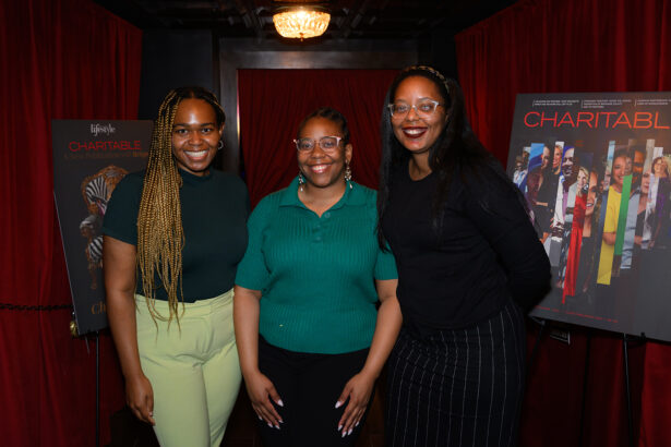Three women stand smiling in front of two posters for “Charitable” in a room with red curtains and a chandelier above. Lifestyle