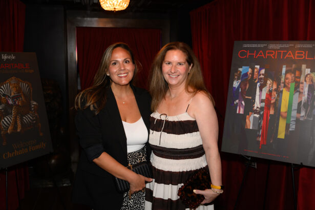 Two women pose and smile at an indoor event with red curtains. On either side of them, posters display the word “CHARITABLE” and photos from the event. Both women wear patterned dresses and stand close together. Lifestyle