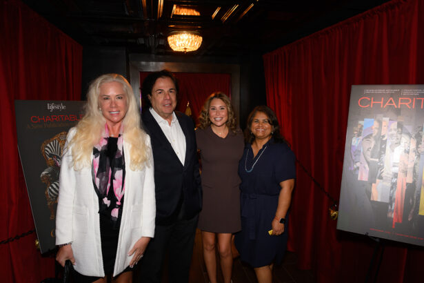Four people stand together, smiling, between two posters at an indoor event with red curtains and dim lighting. The group includes three women and one man in formal attire. Lifestyle