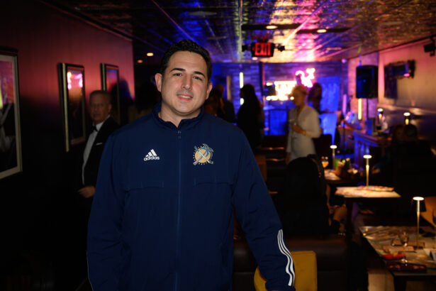 A man in a blue zip-up jacket stands in a dimly lit lounge with people and neon lights in the background. Lifestyle