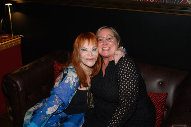 Two women sit closely on a brown leather sofa, smiling at the camera. One wears a blue floral shawl, the other a black dress. The atmosphere is warm and cozy, with dark walls and decorative accents visible. Lifestyle
