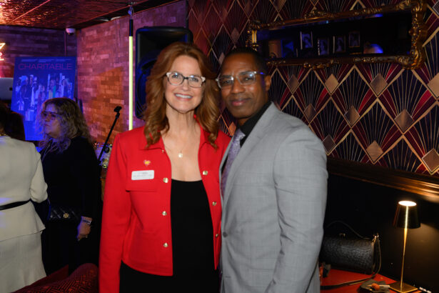 A woman in a red jacket and a man in a gray suit smile together at an indoor event, with patterned wallpaper, framed photos, and people in the background. Lifestyle
