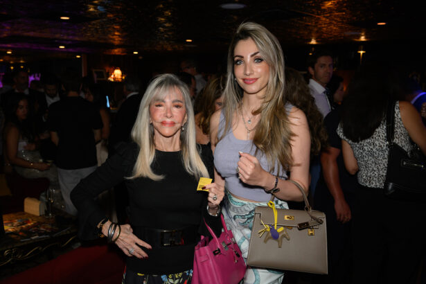 Two women pose and smile at a crowded indoor event. One wears a black top and holds a pink purse, the other wears a light blue outfit and holds a beige handbag. People mingle in the dimly lit background. Lifestyle