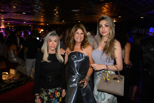 Three women pose together at a lively, upscale event. Two smile at the camera, one holds a wine glass, and the third carries a beige handbag. The background features a crowd in formal attire and colorful lighting. Lifestyle