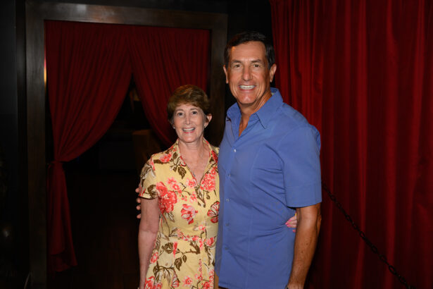 A smiling man in a blue shirt stands beside a smiling woman in a floral dress, both posing in front of red curtains in a dimly lit room. Lifestyle