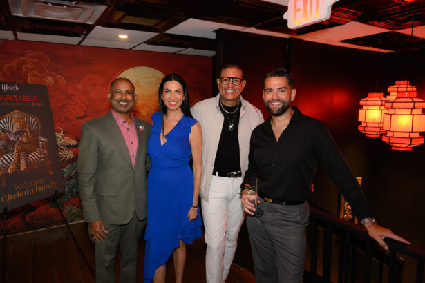 Four people pose together at an indoor event, dressed in formal attire. Warm lighting and decorative lamps illuminate the space. A sign in the background reads “Welcome Chelada Family.”. Lifestyle