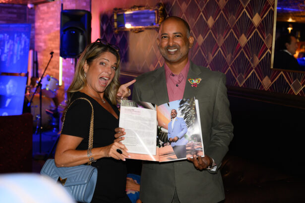 A smiling woman points at a magazine held open by a smiling man in a suit at an indoor event with patterned wallpaper and dim lighting. Lifestyle