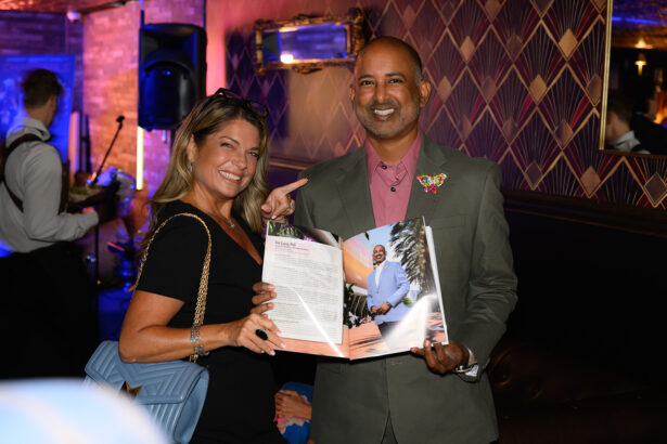A smiling woman and man stand indoors. The man holds open a magazine featuring his own photo, while the woman points at the page. Both are dressed formally and seem happy. Lifestyle