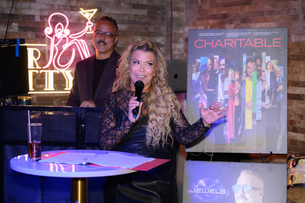 A woman speaks into a microphone at a podium with papers, beside a large “Charitable” poster. A man stands behind her near a keyboard. Neon and brick walls are in the background. Lifestyle
