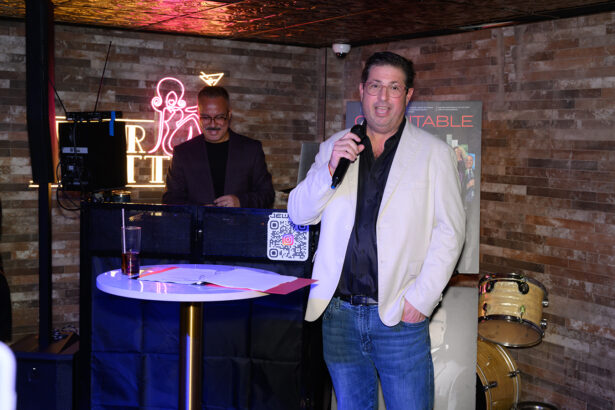 A man in a light jacket speaks into a microphone at an event, with a DJ behind him and a drum set in the background. Neon and brick wall decor are visible. Lifestyle
