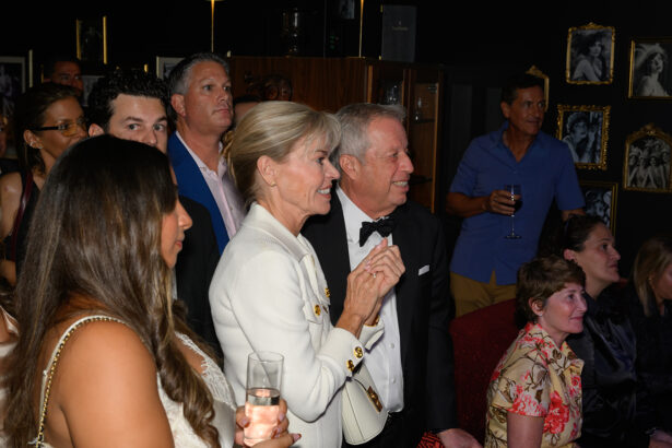 A group of well-dressed people gathers indoors at an event, watching something off-camera. Many are smiling and holding drinks. Framed photos decorate the dark walls behind them. Lifestyle