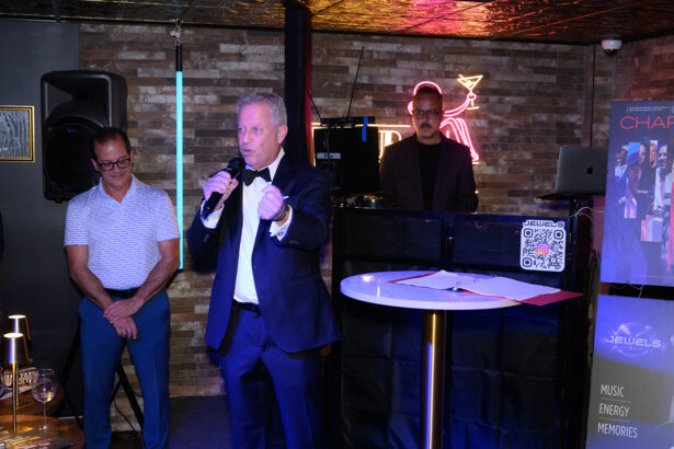 A man in a tuxedo speaks into a microphone at a small indoor event, with two other men standing nearby. A neon sign and DJ booth are visible in the background, and event signage appears on the right. Lifestyle
