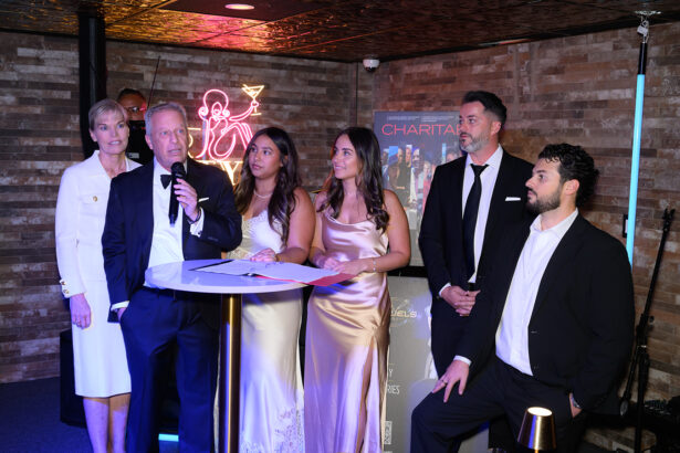 Six people in formal attire stand around a small round table. One man speaks into a microphone. Two women in satin dresses stand in the center. A neon cocktail sign and posters are visible on the brick wall behind them. Lifestyle