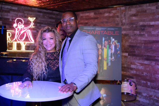 A woman and a man stand together, smiling, at a round table in a dimly lit venue with neon and poster displays in the background. Lifestyle