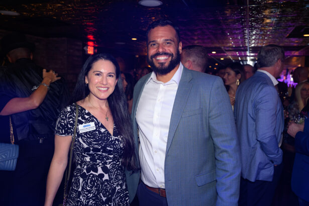 Two people smiling and posing for a photo at a crowded indoor event, dressed in formal attire. Other attendees are visible in the background, engaged in conversation. Lifestyle
