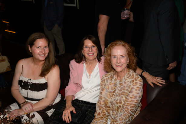 Three women sit close together on a brown sofa at an indoor event, smiling at the camera. The women wear patterned and light-colored outfits. Other people are visible standing in the background, mostly out of focus. Lifestyle