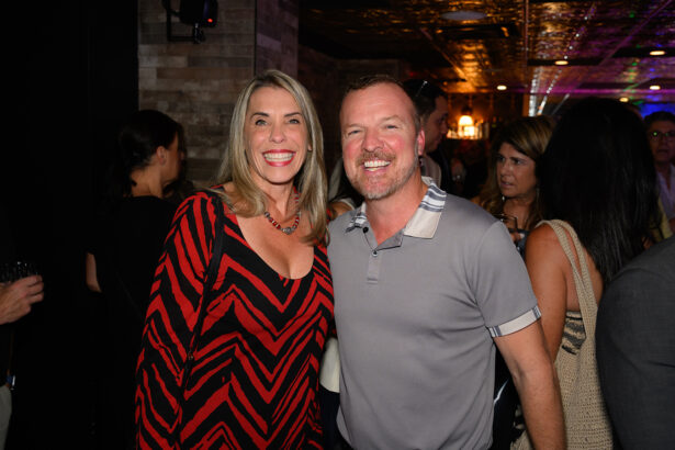 A smiling woman in a red and black dress stands beside a smiling man in a gray shirt at a lively indoor event with people in the background. Lifestyle