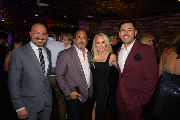 Four people dressed formally are smiling and posing together at an indoor event, with other guests mingling in the background under warm lighting. Lifestyle
