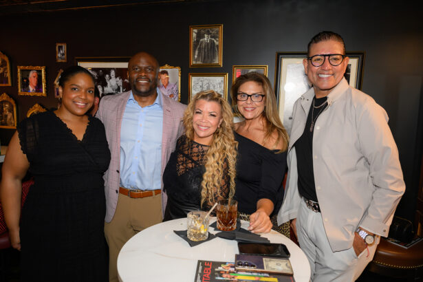 Five adults pose and smile around a small round table with drinks in a warmly lit room decorated with framed photos and art on the wall behind them. Lifestyle