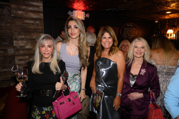 Four women dressed elegantly pose together at a dimly lit indoor event. One holds a wine glass and a pink handbag; another wears a shiny strapless dress. An EXIT sign glows in the background. Other people mingle behind them. Lifestyle