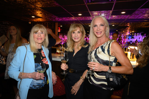 Three women dressed elegantly stand together at a bar, each holding a glass of white wine. The background features a lit bar, ceiling tiles, and other people socializing. All three women are smiling at the camera. Lifestyle