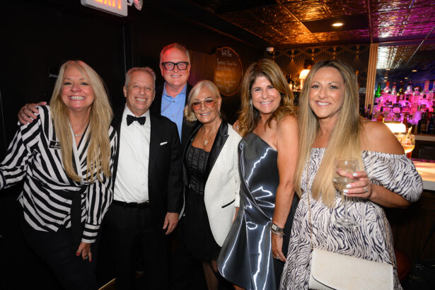 Six adults dressed formally are smiling and posing together in a dimly lit bar or restaurant. One woman holds a wine glass. The background features bar shelves with bottles and warm lighting. Lifestyle